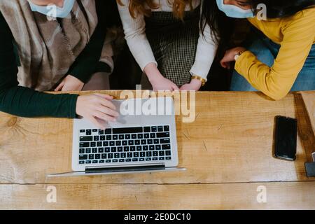 From above group of cropped unrecognizable multiethnic female freelancers in masks working on laptop in cafe while discussing new business project dur Stock Photo