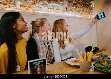 Group of cheerful diverse female friends sitting at table in cafe and taking selfie on smartphone while spending weekend together Stock Photo