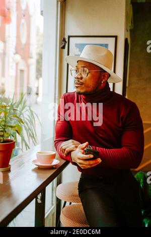Pensive African American male in stylish outfit sitting at counter in coffee shop while chilling at weekend and looking out of window Stock Photo