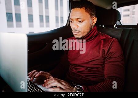 Focused African American male entrepreneur sitting in car on backseat and browsing netbook while working remotely on project Stock Photo