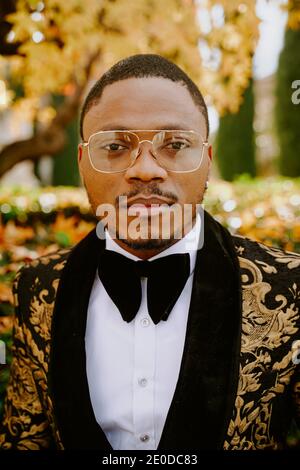 Elegant African American male wearing expensive tuxedo walking along street and looking at camera Stock Photo