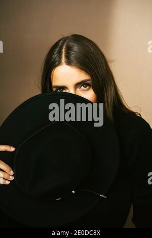 Charming young Woman with long dark hair hiding face behind black hat and looking at camera against beige background Stock Photo