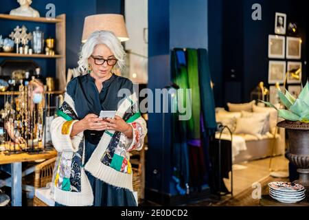 Elegant middle aged female in stylish outfit and glasses messaging on mobile phone while spending time at home with vintage interior Stock Photo