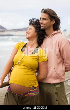 Young pregnant woman leans on long hair husband enjoying sea views in Gran Canaria. Beautiful couple on casual clothing standing by beach walk railing Stock Photo