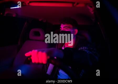 Unrecognizable concentrated young male in casual clothes and face mask driving modern car with glowing neon lights at night during coronavirus Stock Photo