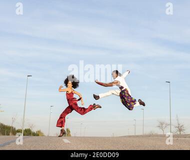 Side view of African American couple in trendy traditional clothes jumping high above asphalt road and performing dance on background of blue sky Stock Photo