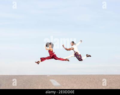 Side view of African American couple in trendy traditional clothes jumping high above asphalt road and performing dance on background of blue sky Stock Photo