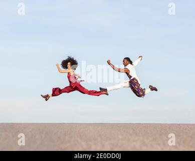 Side view of African American couple in trendy traditional clothes jumping high above asphalt road and performing dance on background of blue sky Stock Photo