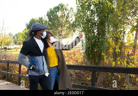 Stylish African American couple in medical masks hugging and walking in autumn park during coronavirus epidemic Stock Photo
