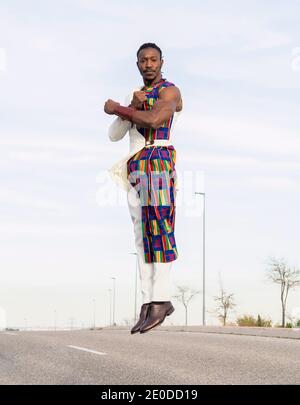 African male dancer in trendy outfit levitating above road while performing energetic movements against blue sky making arms gesture looking at camera Stock Photo
