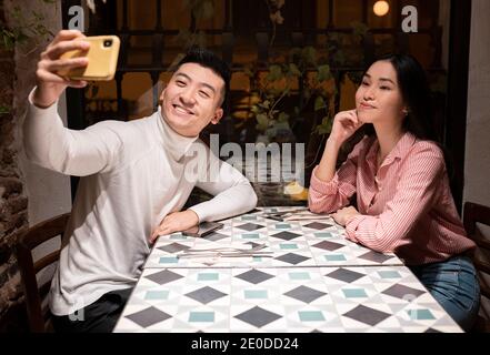 Side view of cheerful young Asian man and Woman in casual clothes sitting at table and taking selfie on smartphone while having meeting in cozy cafe Stock Photo