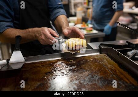Crop anonymous cook with spatula taking roasted meat cutlet with melted cheese while preparing hamburger in restaurant kitchen Stock Photo
