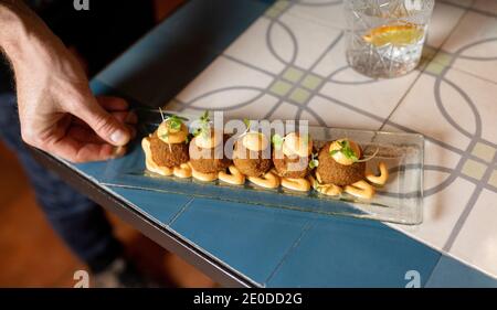 From above of crop anonymous waiter putting tray with delicious deep fried croquettes garnished with cream cheese and herbs on table while serving foo Stock Photo