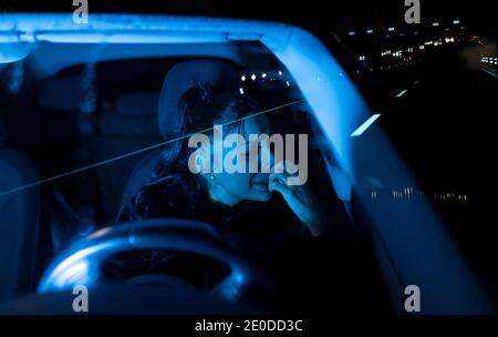 Through windshield view of upset young female driver crying about personal problems while sitting in car parked on night road Stock Photo