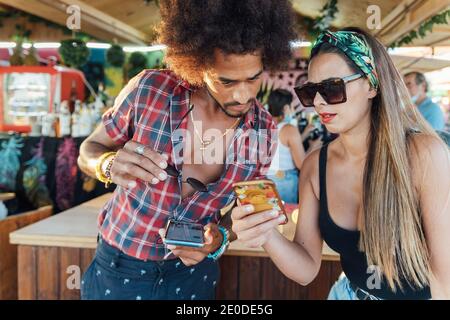 African American man showing photo on mobile phone to girlfriend while standing near counter of outdoor beach bar Stock Photo