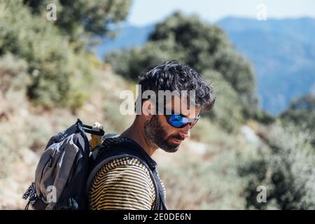 Side view of adult bearded male mountaineer in sunglasses and with backpack standing against blurred background of rocky highland Stock Photo