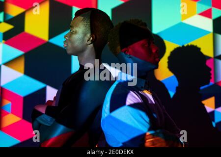 Side view of emotionless African American men standing back to back in studio with colorful neon illumination in shape of cubes Stock Photo