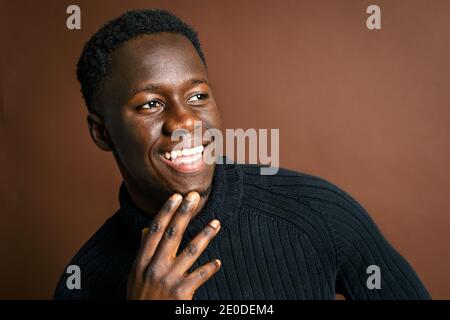 Side view African American male rubbing chin and leaning on hand while standing on brown background in studio and looking away Stock Photo