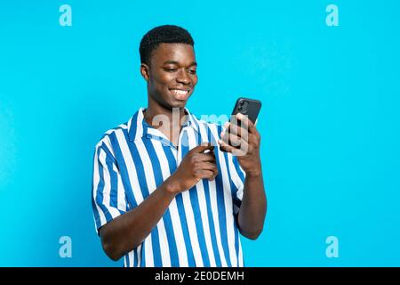 Delighted African American male browsing smartphone and reading good news about business project while standing with clenched fist on vivid blue backg Stock Photo