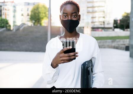 Stylish young African American businessWoman in protective mask standing on city street and messaging on smartphone Stock Photo