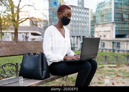 Young African American businessWoman with face mask in elegant clothes sitting on bench and working on laptop against blurred modern urban background Stock Photo