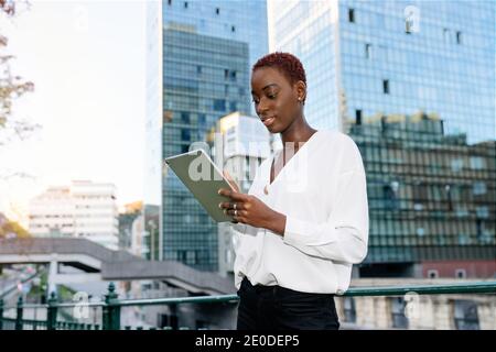 Contemporary African American female entrepreneur in stylish outfit browsing tablet while standing against modern glass building in city Stock Photo