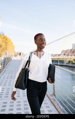 Confident successful positive young black businessWoman with short hairstyle standing near river against modern buildings in downtown Stock Photo