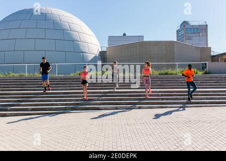 Company of diverse runners running down stairs in city while doing cardio workout together on sunny day in summer Stock Photo