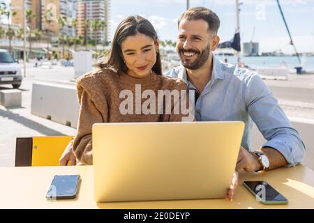 Side view of happy young ethnic smiling and pointing at laptop screen while watching funny video with excited stylish girlfriend on cafe terrace on su Stock Photo