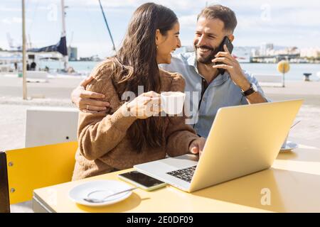 Happy young male talking on smartphone and hugging cheerful ethnic girlfriend drinking coffee and working remotely on laptop in outdoor cafe Stock Photo
