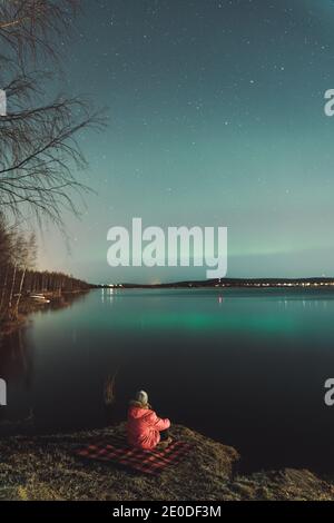 Back view of unrecognizable explorer sitting on shore of lake at night and enjoying spectacular scenery of Northern lights in winter Stock Photo