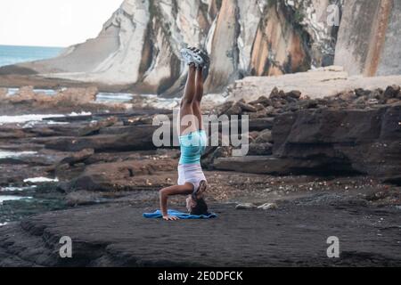 Back view of tranquil female balancing in Salamba Sirsasana on rocky beach while practicing yoga in summer Stock Photo