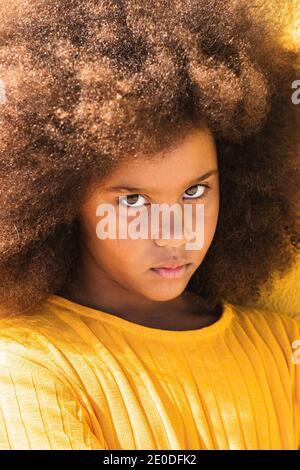 Serious teen African American girl with fluffy curly hair wearing yellow sweatshirt looking at camera Stock Photo