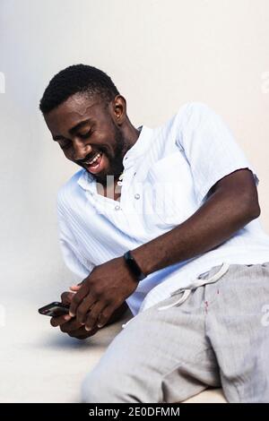 Positive cheerful African American male in casual outfit relaxing using smartphone on white background in studio and laughing Stock Photo