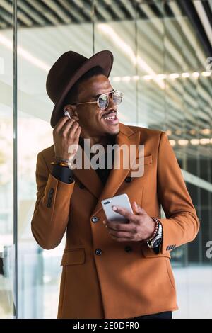 Positive young African American man in stylish suit and hat smiling while having phone conversation using wireless earphones and looking away in moder Stock Photo