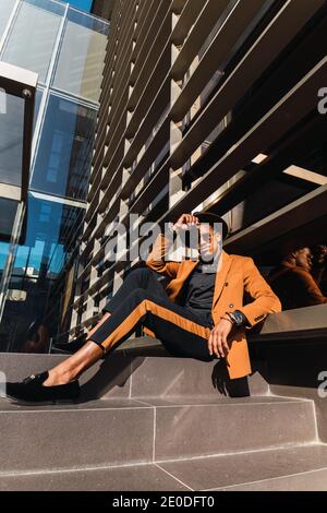 Low angle of stylish young African American businessman in fashionable suit and hat while sitting for meeting near contemporary skyscraper Stock Photo
