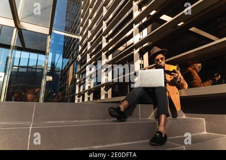 Serious African American male wearing trendy outfit and accessories sitting in street with laptop and plastic card and making purchase online Stock Photo