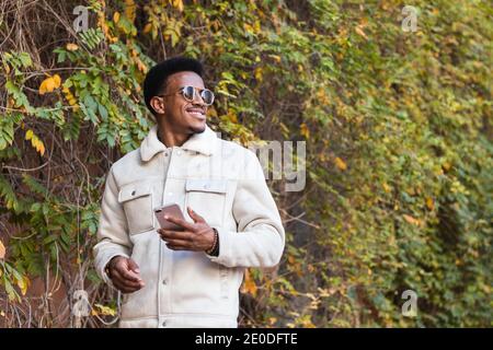 Low angle of happy young African American guy in trendy jacket and sunglasses smiling and looking away while using smartphone in autumn park Stock Photo