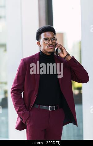 Serious young African American male manager in stylish suit and eyeglasses discussing business issues on smartphone and looking away Stock Photo
