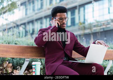 Pensive young African American male freelancer in stylish outfit and eyeglasses sitting on bench in city park and having phone conversation during rem Stock Photo