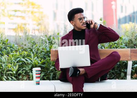 Pensive young African American male freelancer in stylish outfit and eyeglasses sitting on bench in city park and having phone conversation during rem Stock Photo