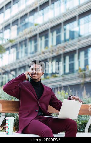 Happy young African American male freelancer in stylish outfit and eyeglasses sitting on bench in city park and having phone conversation during remot Stock Photo