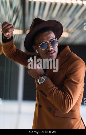 Side view of young handsome black guy in trendy suit and sunglasses while standing in modern building hall with headphone Stock Photo