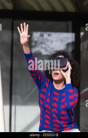 Excited female wearing modern VR glasses experiencing virtual reality while riding roundabout on playground with raised arms Stock Photo