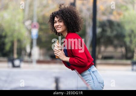 Side view of young African American female standing on street with fresh juice in plastic takeaway cup and enjoying weekend in city Stock Photo