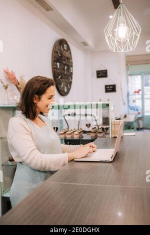 Smiling adult female entrepreneur standing at counter and using laptop while working in cozy cafe Stock Photo