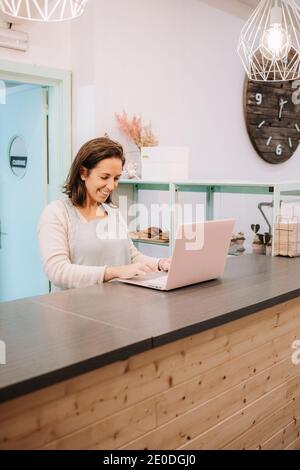 Smiling adult female entrepreneur standing at counter and using laptop while working in cozy cafe Stock Photo
