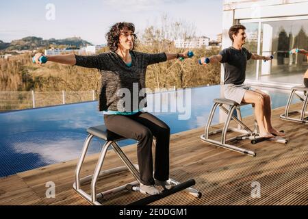 Content people sitting on pilates chairs and practicing with dumbbells while smiling and looking away Stock Photo