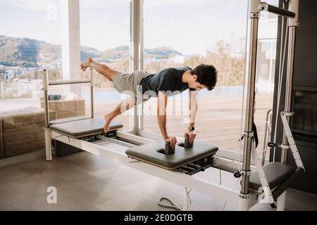 Side view of concentrated male balancing in Eka Pada Phalakasana on pilates machine while doing yoga and practicing mindfulness Stock Photo