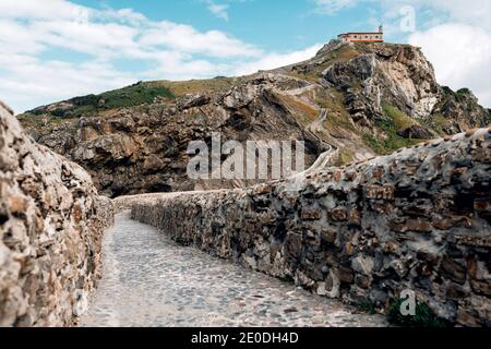 Empty road leading along stone bridge and ridge of rocky hill to lonely country house on island Gaztelugatxe surrounded by tranquil sea water with whi Stock Photo